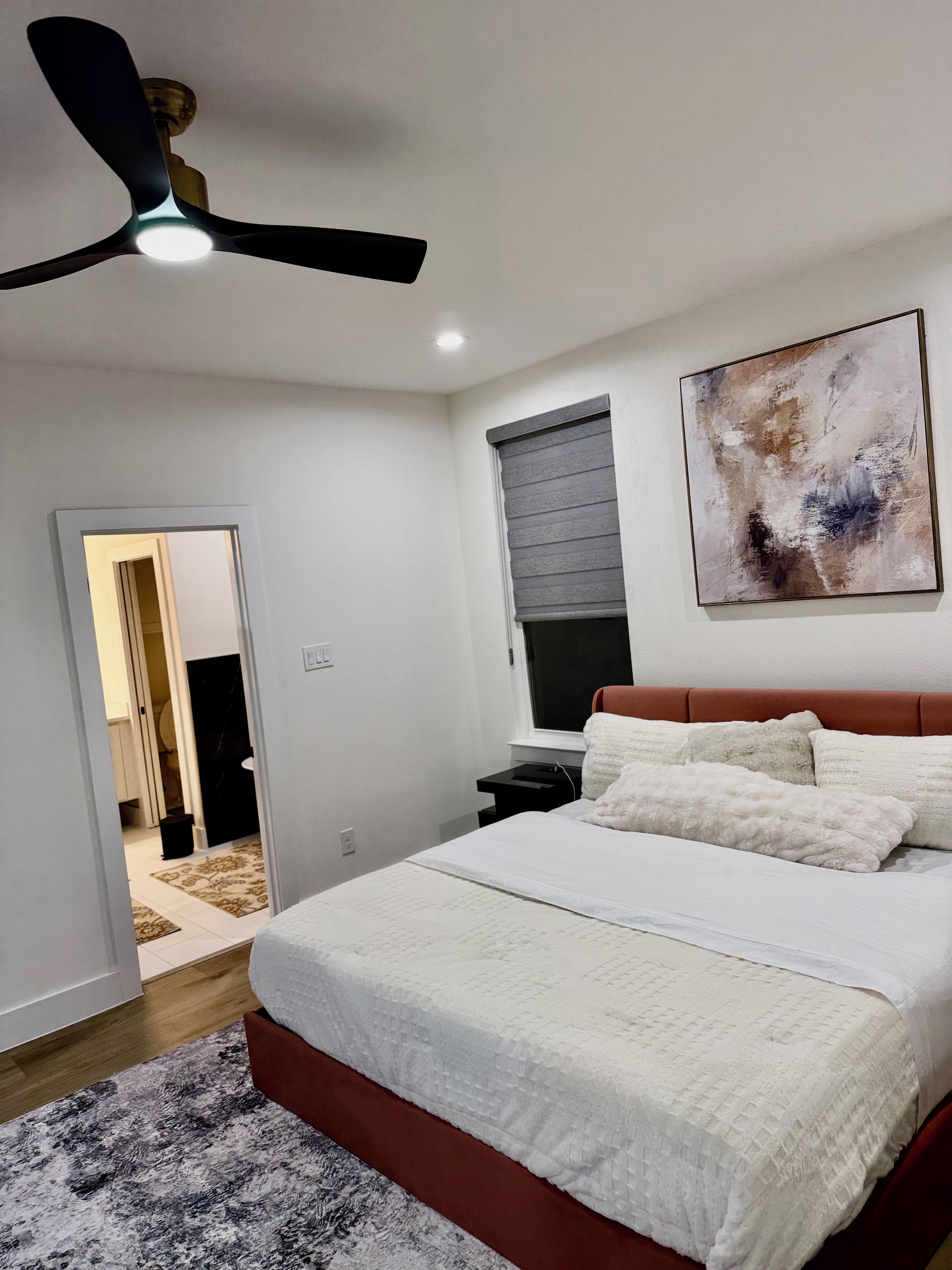 A bedroom with a bed, a nightstand, a ceiling fan, a window with blinds, and a painting on the wall.