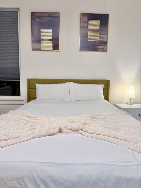 A hotel room with a bed, two wall-mounted artworks, a window with blinds, and a bedside lamp.