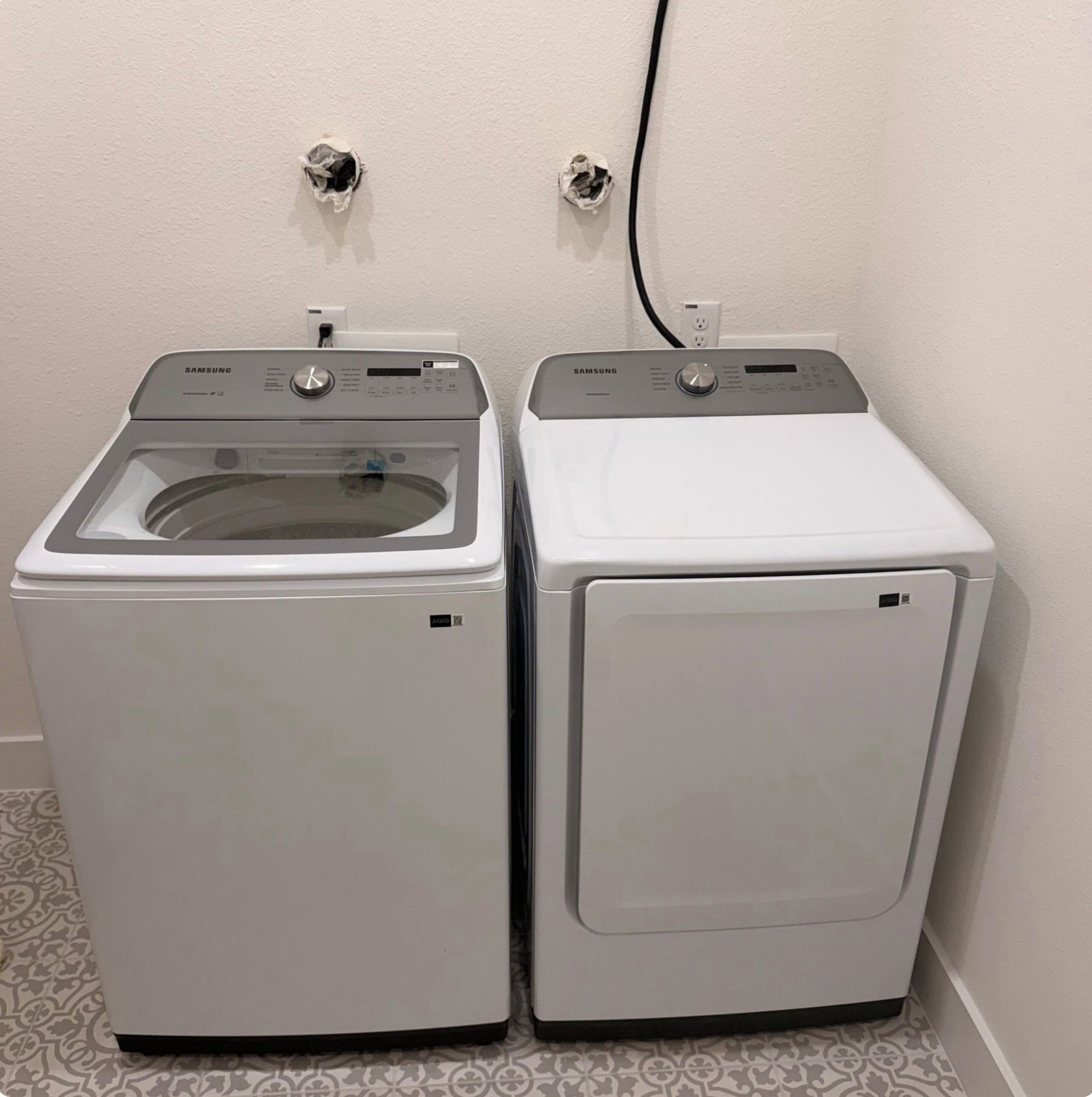 Two white front-loading washing machines side by side with a power outlet and cable on the wall.