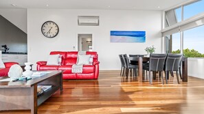 Ein modernes Wohnzimmer mit einem roten Ledersofa, einem Essbereich mit schwarzen Stühlen, einem Holztisch, einer Wanduhr und einem Gemälde vom Strand.