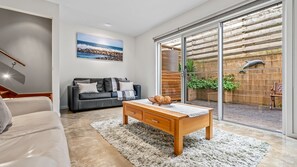 A modern living room with a sofa, coffee table, and a large sliding glass door leading to a patio with a wooden fence and a dolphin sculpture. - King Tide Apartment - 55 Philip (Port Fairy)
