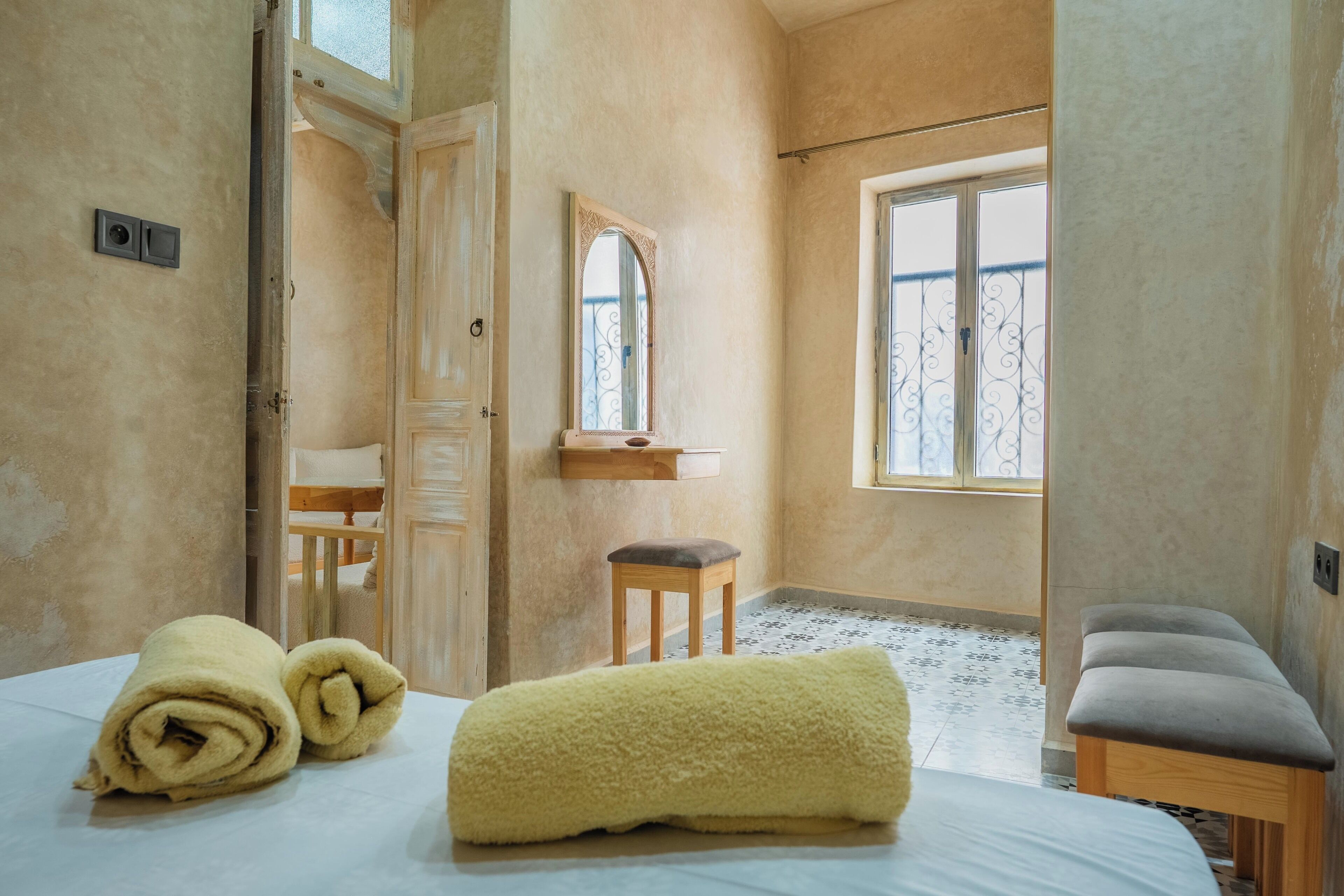 A room with a bed, a bench, a window, and a mirror.