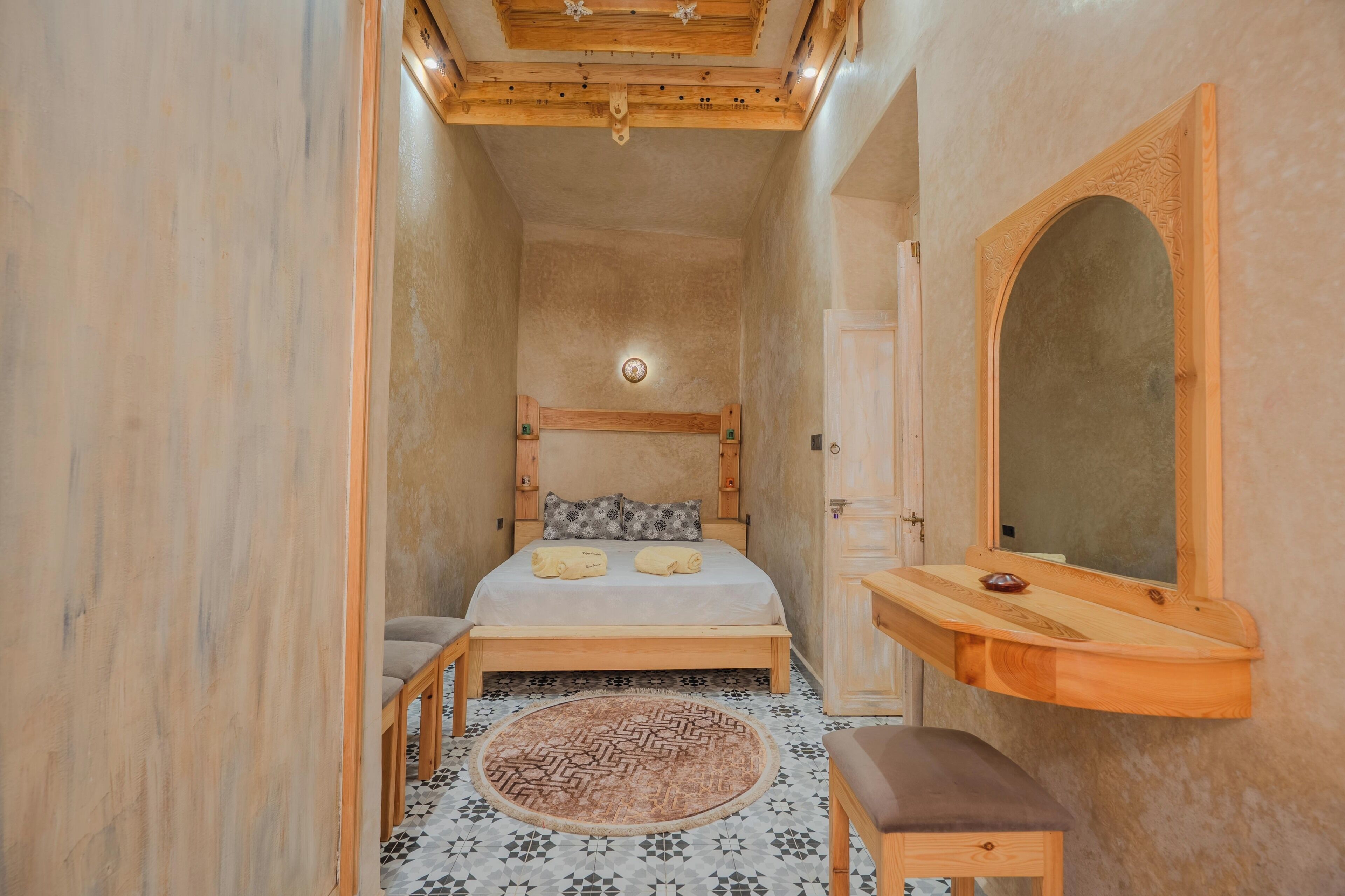 Una habitación estrecha con cama, un banco de madera, un tapete redondo y un espejo.