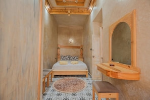 Een kleine kamer met een bed, een houten bankje, een rond tapijt en een spiegel.