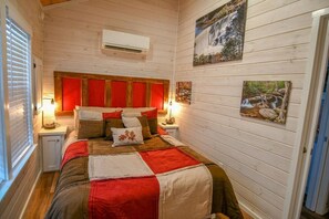 Ein gemütliches Schlafzimmer mit Holzboden, einem Bett mit rot-beiger Bettwäsche, zwei Nachttischen mit Lampen und Wandbildern.