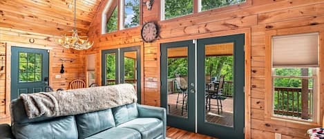 Ein Interieur einer Holzhütte mit einem Ledersofa, einem Kronleuchter und einem Essbereich, der durch Glastüren einsehbar ist.
