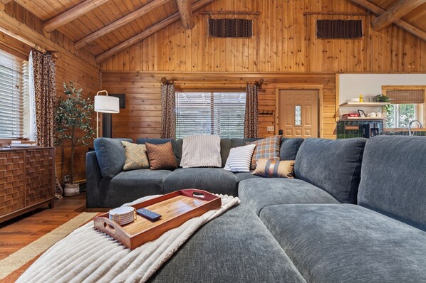 A cozy living room with a sectional sofa, a wooden ceiling, and a kitchen area visible through an open door.