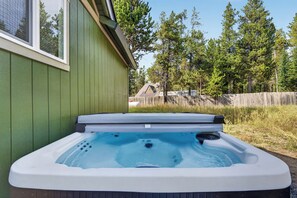 A hot tub in front of a green building with a wooden fence and trees in the background.