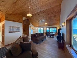 A spacious living area with a wooden ceiling, a sofa, a dining table with chairs, and a view through a large window. - Crane Cabin-Set directly along the sandy shores of Amnicon Bay, Aajijash (La Pointe)