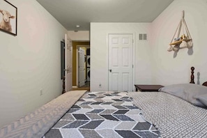 A bedroom with a bed, a bedside table, a hanging light fixture, and a door leading to another room.