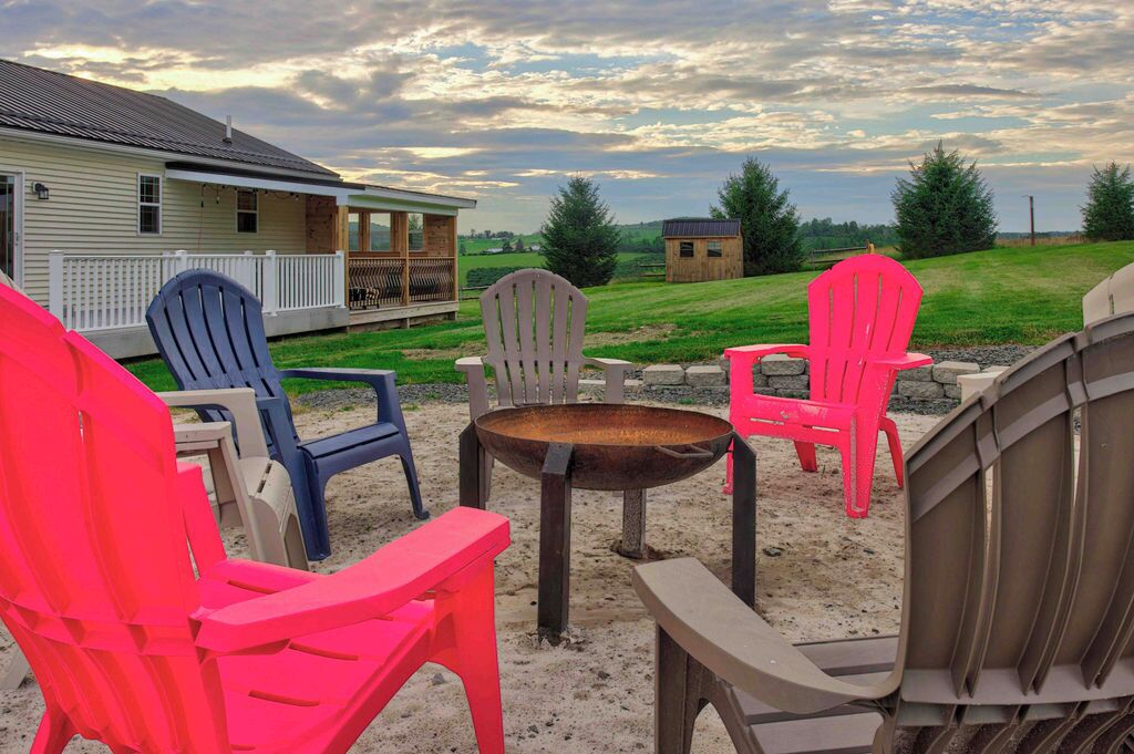 Une terrasse dotée de chaises Adirondack et d’un foyer, offrant une vue sur une zone herbeuse avec des arbres et une maison en arrière-plan.