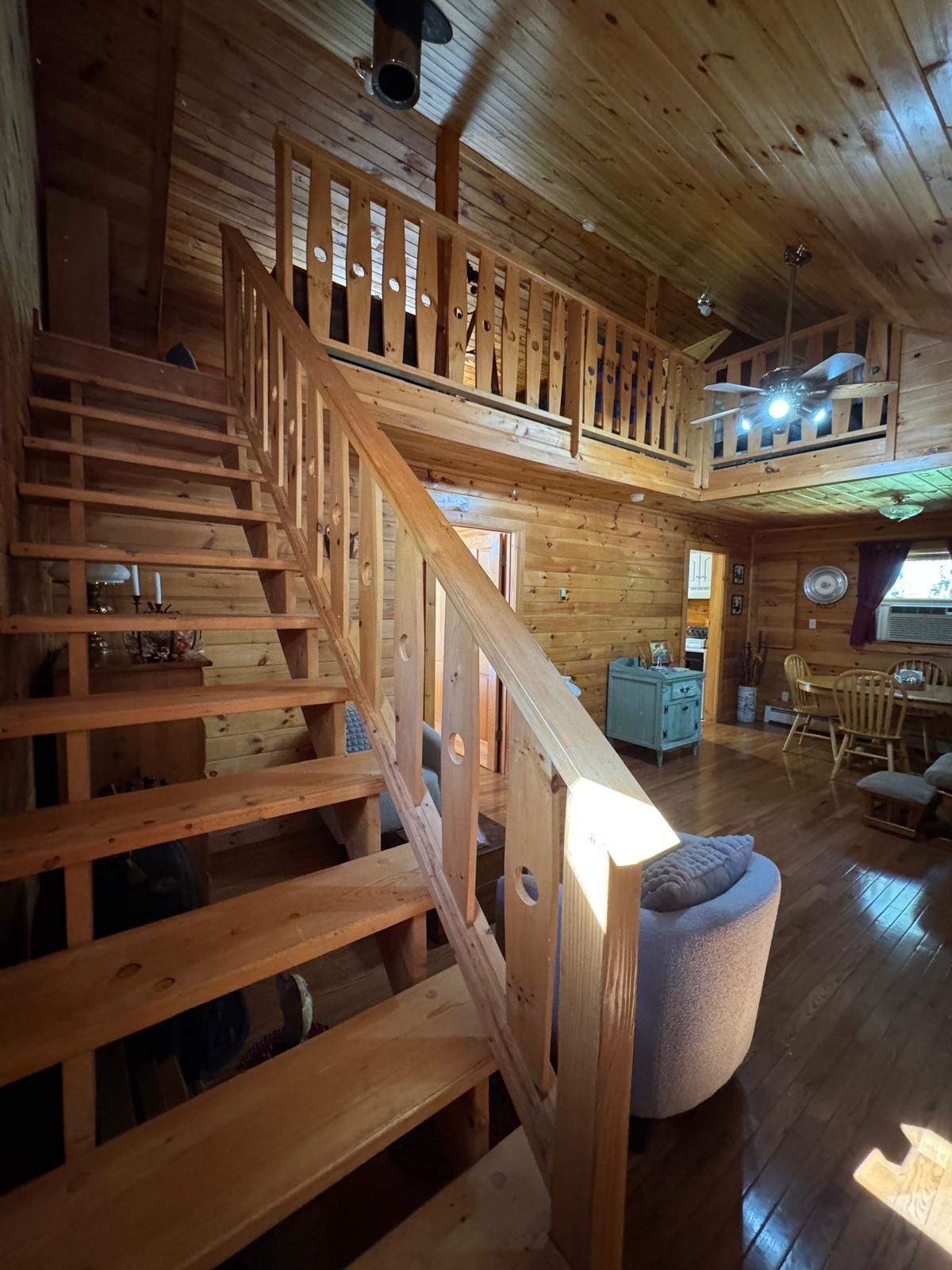 A wooden interior with a staircase leading to an upper level, a dining area, and a seating area with a round ottoman.