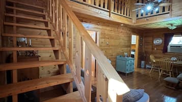 A wooden interior with a staircase leading to an upper level, a dining area, and a seating area with a round ottoman.