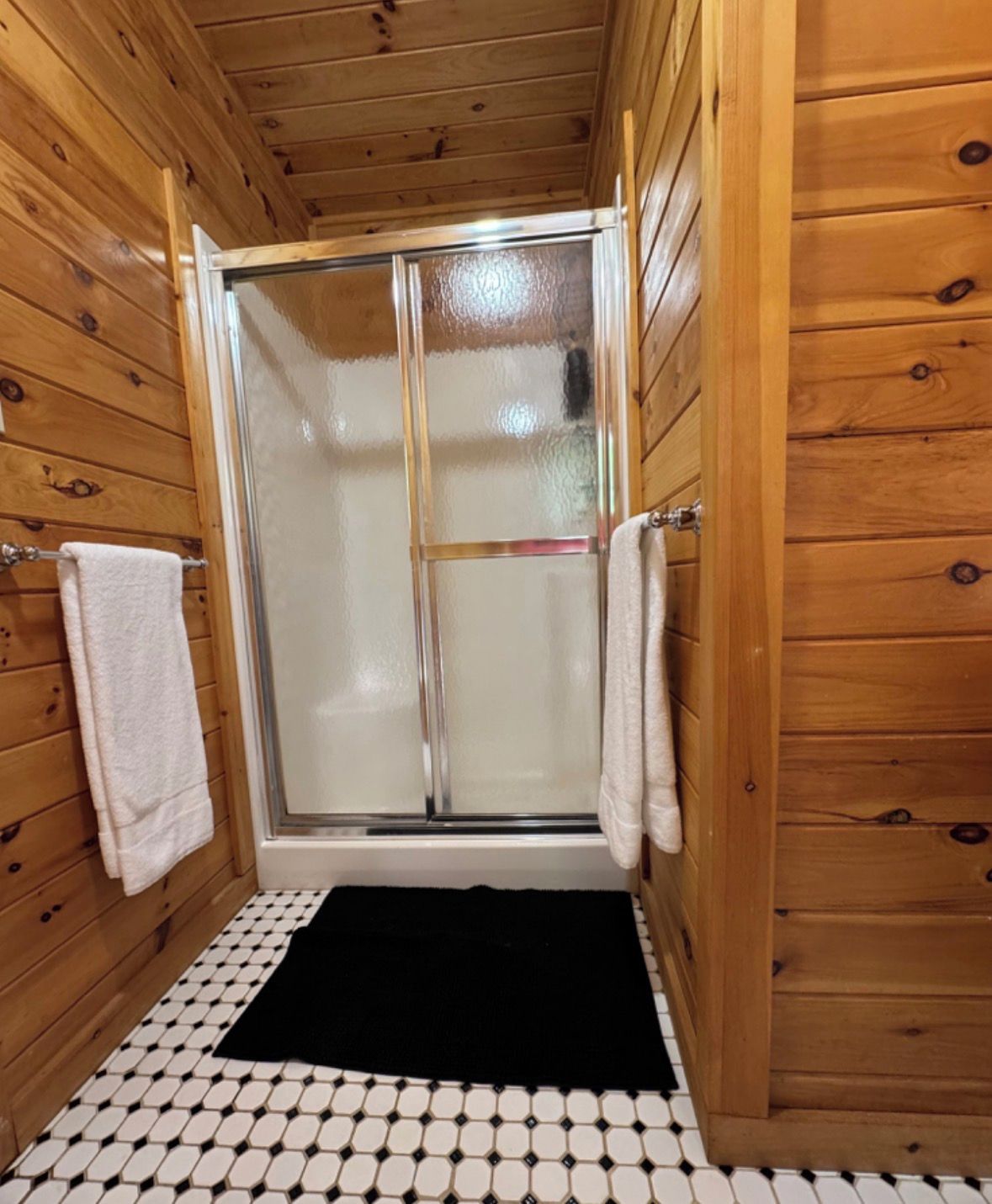 A wooden cabin bathroom with a glass shower enclosure, black and white patterned floor, and white towels on hooks.