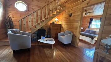 A wooden cabin interior with a staircase, two armchairs, a table with a vase, and a bed in the adjacent room.