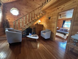 Interior de una cabaña de madera con escalera, dos sillones, una mesa con un florero y una cama en la habitación contigua.