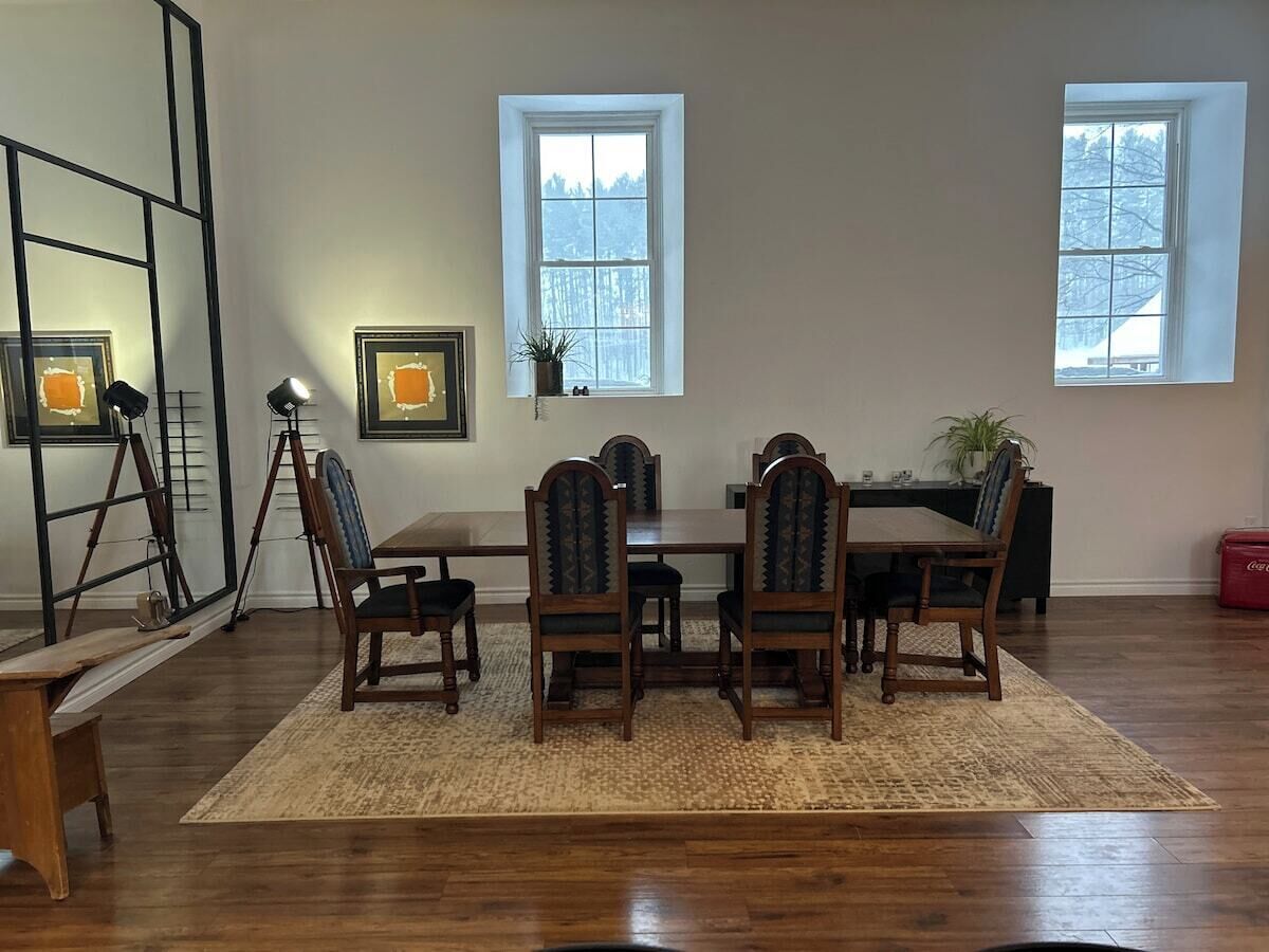 A dining area with a wooden table and chairs, two windows, and a red cooler.