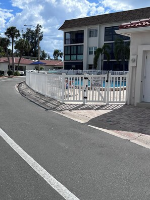 A gated pool area with a white fence, a clear sky, and a building with multiple windows in the background.