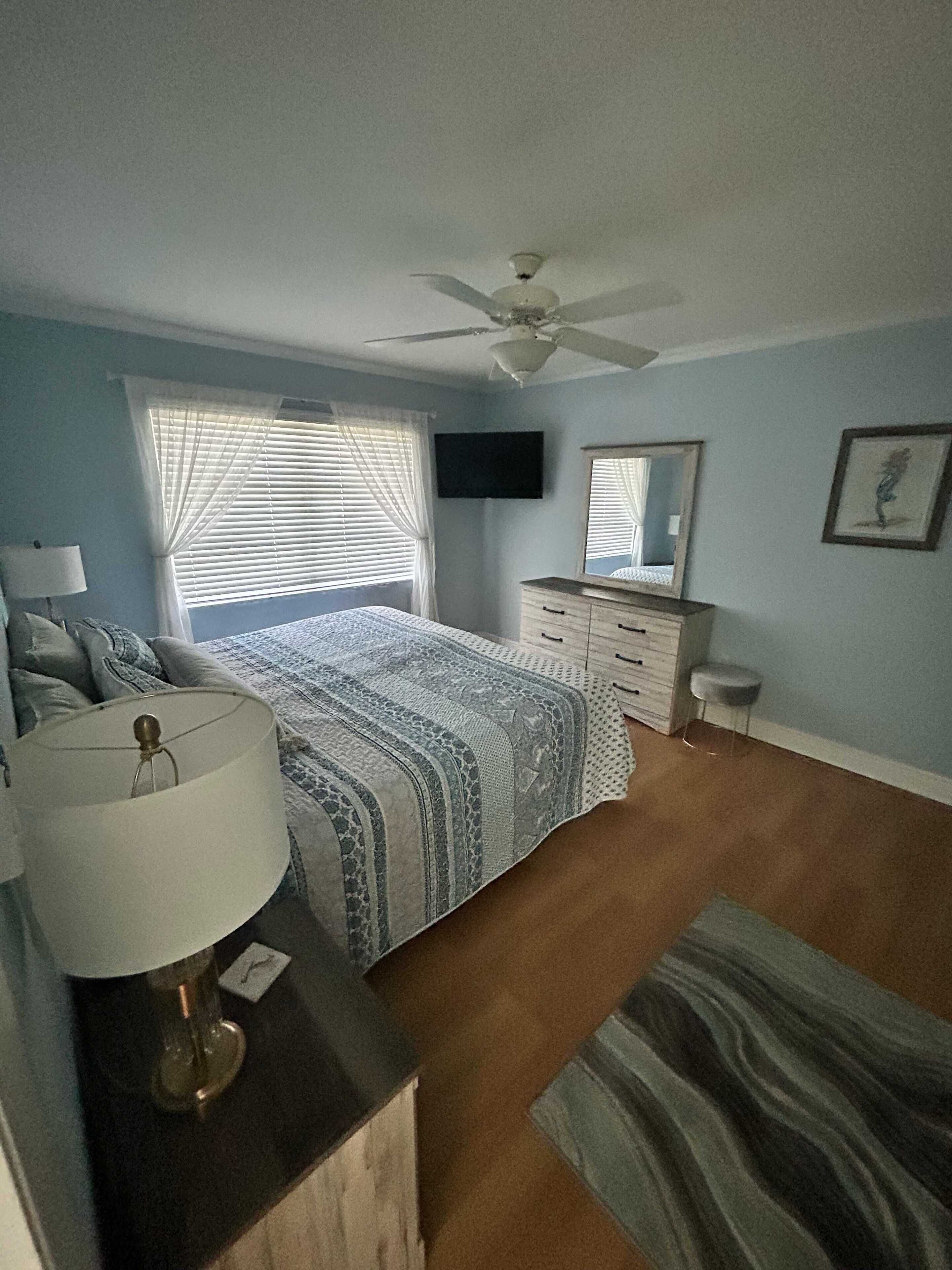 A bedroom with a bed, a ceiling fan, a TV, a dresser, a mirror, and a window with blinds.