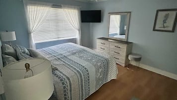 A bedroom with a bed, a ceiling fan, a TV, a dresser, a mirror, and a window with blinds.