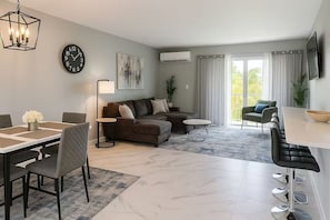 A modern living room with a sofa, dining table, and chairs, a clock, and an air conditioning unit. - Enchanting 1-bedroom condo with AC in vibrant Freeport (Freeport)