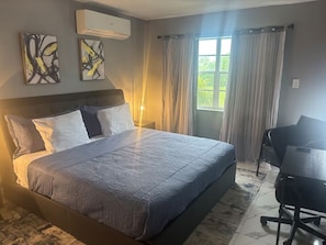 A bedroom with a bed, a desk, and a chair. There is a window with curtains, an air conditioning unit, and two abstract paintings on the wall. - Enchanting 1-bedroom condo with AC in vibrant Freeport (Freeport)