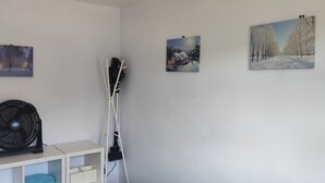 Ein Zimmer mit weißer Wand, einem Standventilator, einer weißen Kommode mit Schubladen und drei gerahmten Bildern, die Winterlandschaften zeigen.