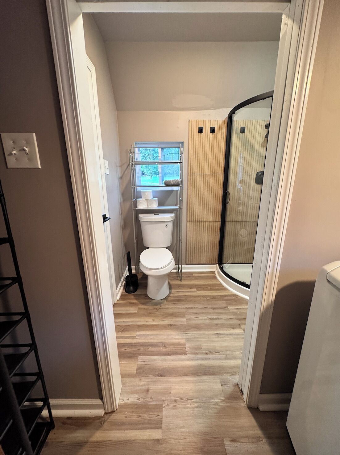 Un baño con inodoro, repisas con artículos de tocador, zona de ducha y piso de madera.