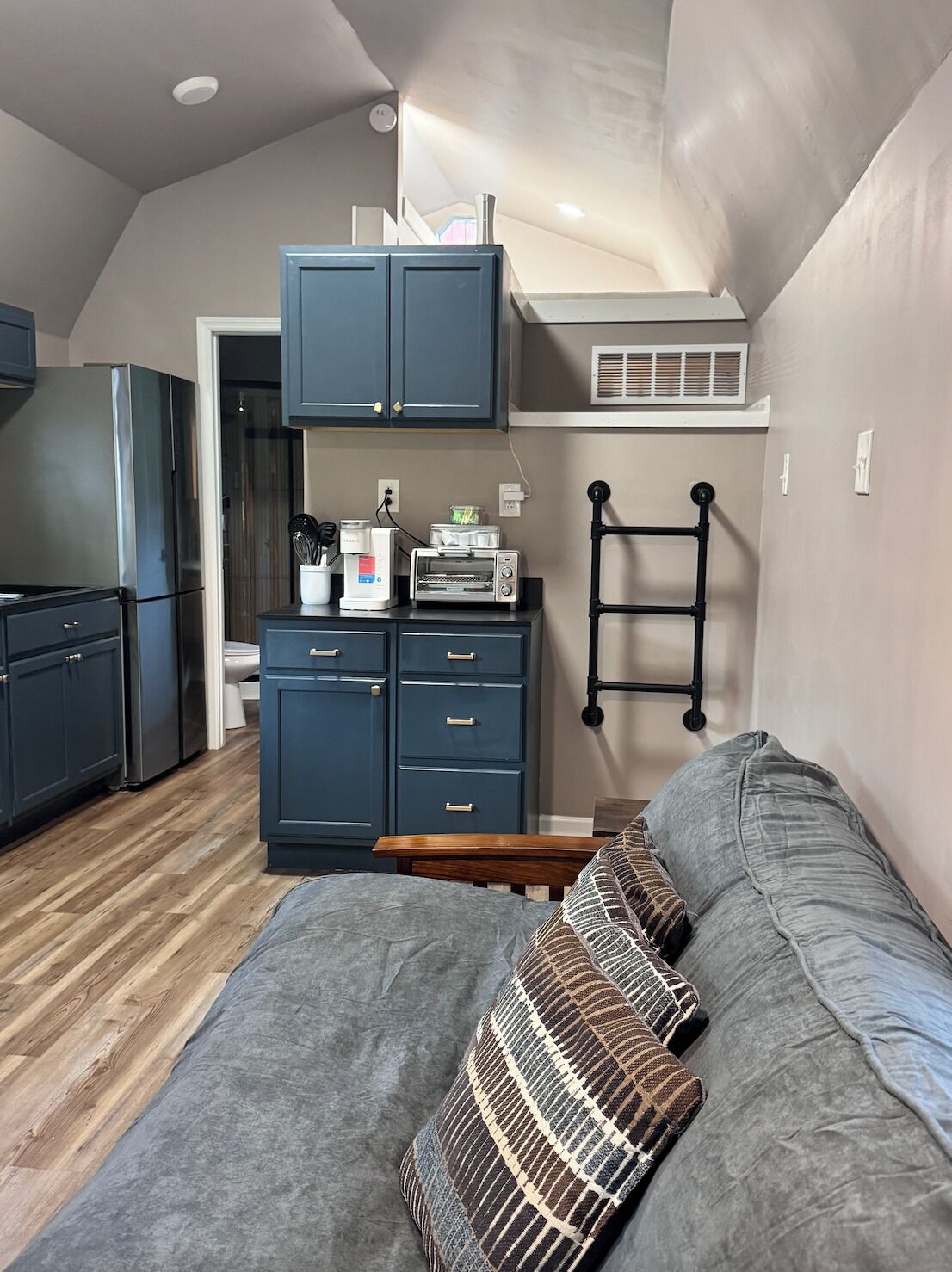 A compact living space with a kitchenette, a bed with patterned pillows, and a built-in shelf above the bed.