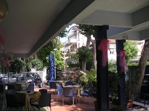 An outdoor seating area with tables and chairs, a blue sign with 'HOTEL' and 'CANASTA' text, and a building in the background. - Canasta (Riccione)