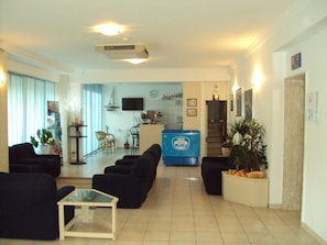 A living room with sofas, a coffee table, and a TV. There is a kitchenette in the background with a refrigerator and a sink. - Canasta (Riccione)