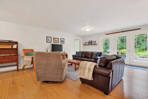 A living room with a brown leather sofa, a wooden coffee table, a brown armchair, a wooden desk, and a television. There are framed pictures on the wall, and a window with a view of trees. - 3BR Bethel retreat with patio, fire pit, WiFi, grill and parking. (Bethel)