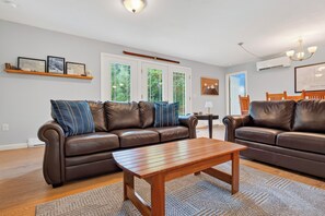 A modern living room with two leather sofas, a wooden coffee table, and a large window with a view of greenery. - 3BR Bethel retreat with patio, fire pit, WiFi, grill and parking. (Bethel)
