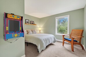 A bedroom with a bed, a chair, a window, and a wall-mounted arcade game. - 3BR Bethel retreat with patio, fire pit, WiFi, grill and parking. (Bethel)
