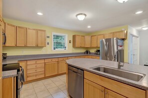 A kitchen with wooden cabinets, a stainless steel refrigerator, a dishwasher, and a window with a view of trees. - 3BR Bethel retreat with patio, fire pit, WiFi, grill and parking. (Bethel)
