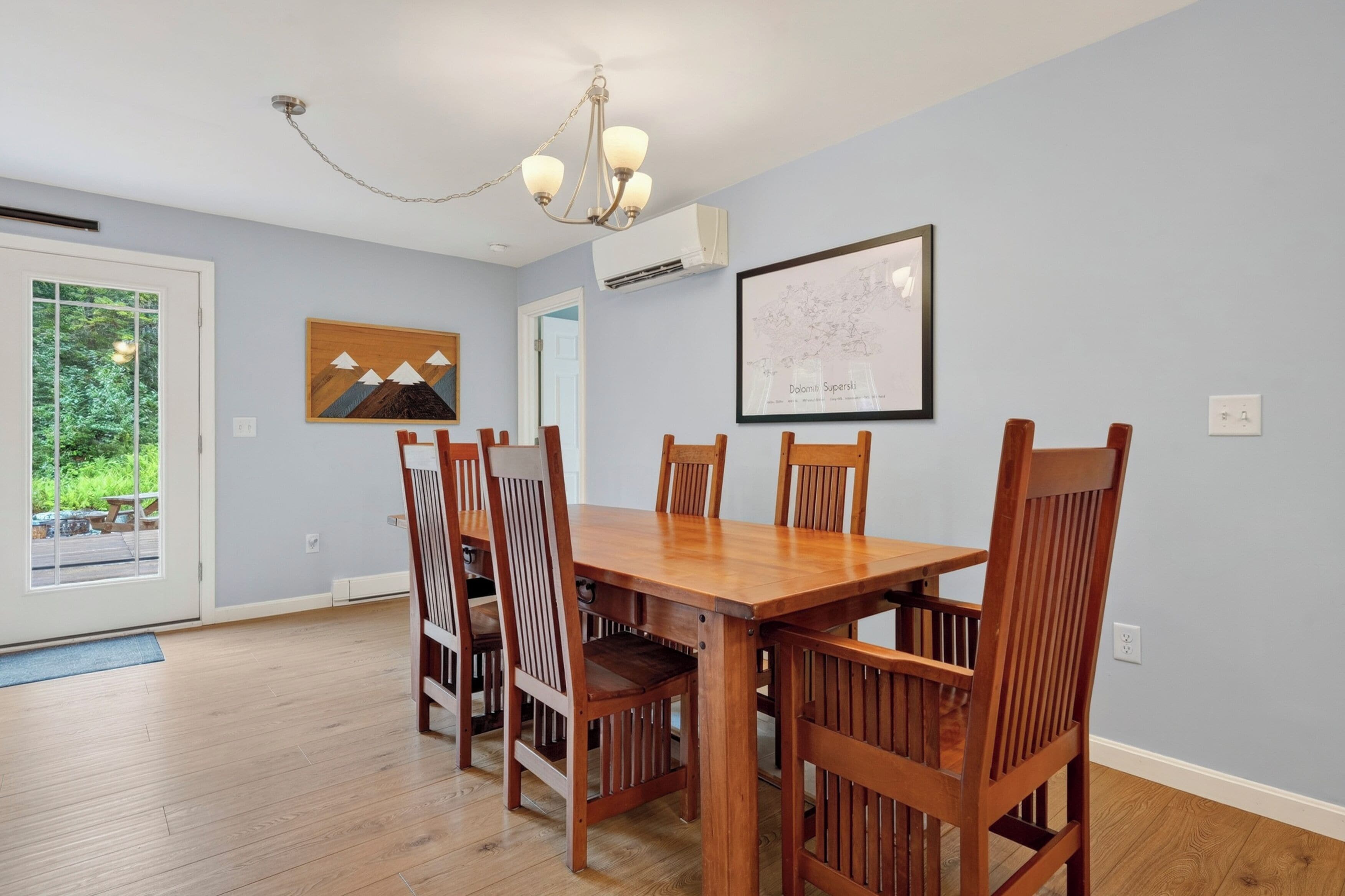 Uma sala de jantar com mesa e cadeiras de madeira, um candelabro, um mapa emoldurado na parede e uma porta que dá para uma varanda.