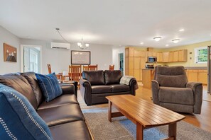 A modern living room with a sofa, two armchairs, and a coffee table. There is a dining area with a table and chairs, and a kitchen with wooden cabinets and appliances. - 3BR Bethel retreat with patio, fire pit, WiFi, grill and parking. (Bethel)