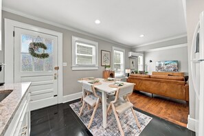 A modern kitchen with a dining area, a living room with a brown leather sofa, and a large window with blinds.