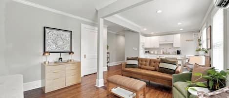 A modern living room with a brown leather sofa, a green armchair, and a wooden bench. There is a kitchen in the background with white cabinets and a dining area.