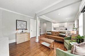 Een moderne woonkamer met een bruine leren bank, een groene fauteuil en een houten bankje. In de achtergrond bevindt zich een keuken met witte kasten en een eethoek.