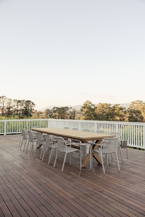 Terraza al aire libre con una mesa y sillas de madera, rodeada por una cerca blanca, con vistas a un paisaje de árboles y colinas.