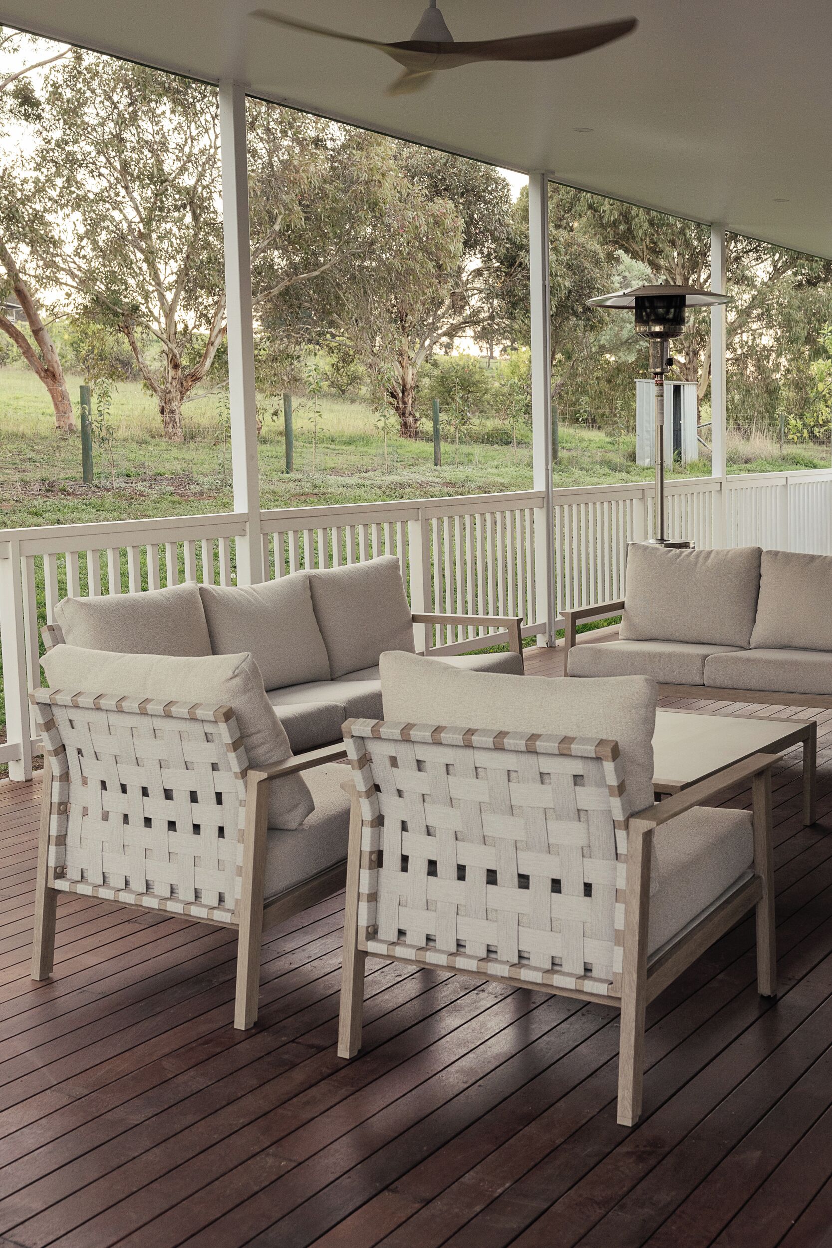 A covered outdoor seating area with a white fence, wooden flooring, and a ceiling fan.