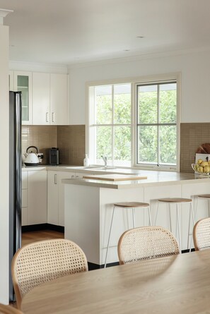 A modern kitchen with white cabinetry, a large window, and a dining area with bar stools. - Yarra Valley Lemon Grove Stay | Fireplace, Mountain Views, Sleeps 8 (Seville)