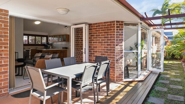 An open-plan living area with a kitchen, dining table, and chairs, connected to a patio with a wooden deck.