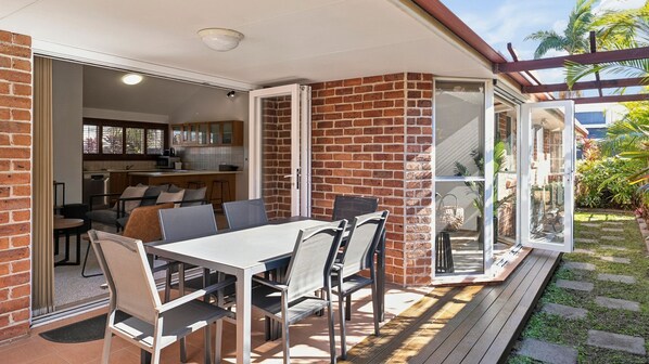 An open-plan living area with a kitchen, dining table, and chairs, connected to a patio with a wooden deck.