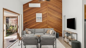 A modern living room with a herringbone wood wall, a grey sofa, two armchairs, a flat-screen TV, and a small side table.