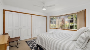 A bedroom with a bed, a desk, a chair, a ceiling fan, and a window with a view of greenery.