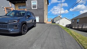 Una calle residencial con una camioneta SUV gris estacionada al costado, casas con diversos estilos arquitectónicos y un cielo despejado.