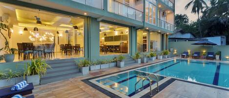 A modern hotel with a swimming pool, outdoor seating, and a well-lit interior.