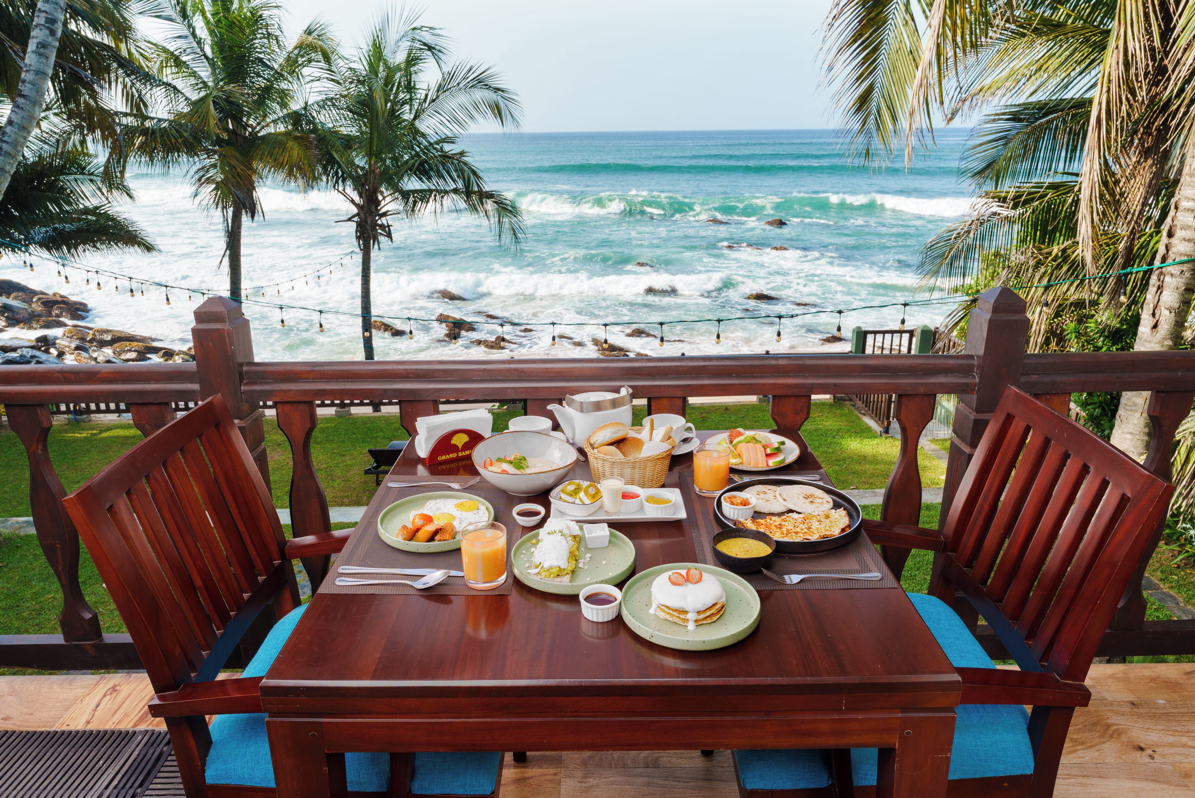 A wooden table set with breakfast items overlooking a beach with waves and palm trees.